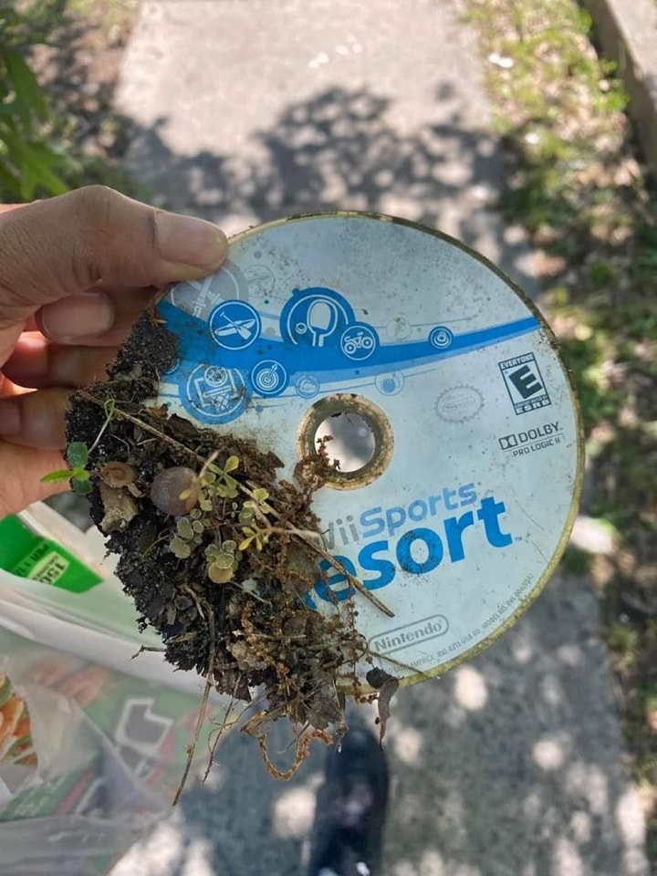 This meme is a photo of a person holding up a dirty, old, and broken Wii Sports Resort game disc, with a small amount of dirt and plants growing out of it. The text on the disc reads "Wii Sports Resort" and has a few scratches and marks on it. The meme is likely poking fun at the idea that even the most high-tech and advanced technology can eventually fall apart and become a part of the natural world.